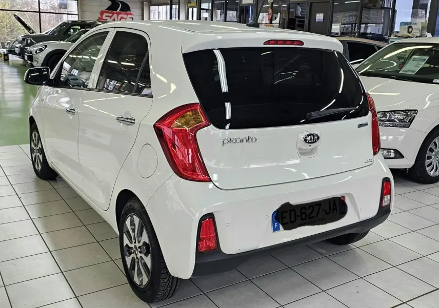 Vue 3/4 arrière droite d'une KIA Picanto blanche 2016 en showroom, avec vitres teintées et feux arrière rouges.
