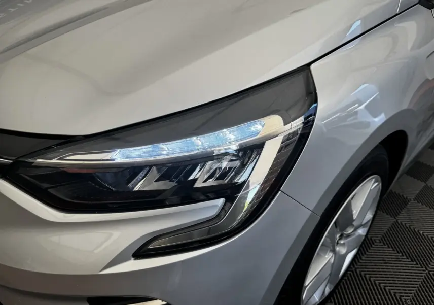 Gros plan sur le phare avant droit allumé de la Renault Clio Business E-TECH 140 gris clair, avec jante visible.