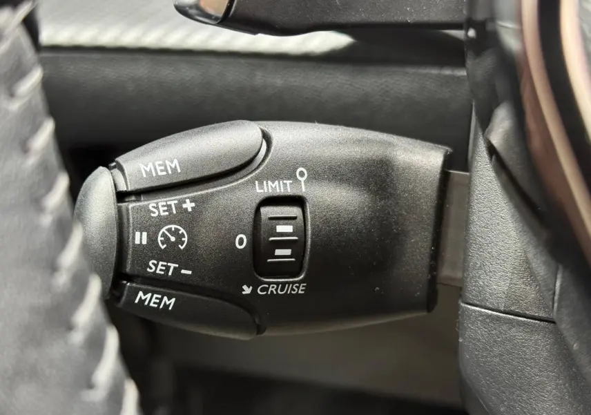 Gros plan sur la commande de régulateur de vitesse à gauche du volant dans l’habitacle noir du Peugeot 208 1.2 Hybrid 2025.