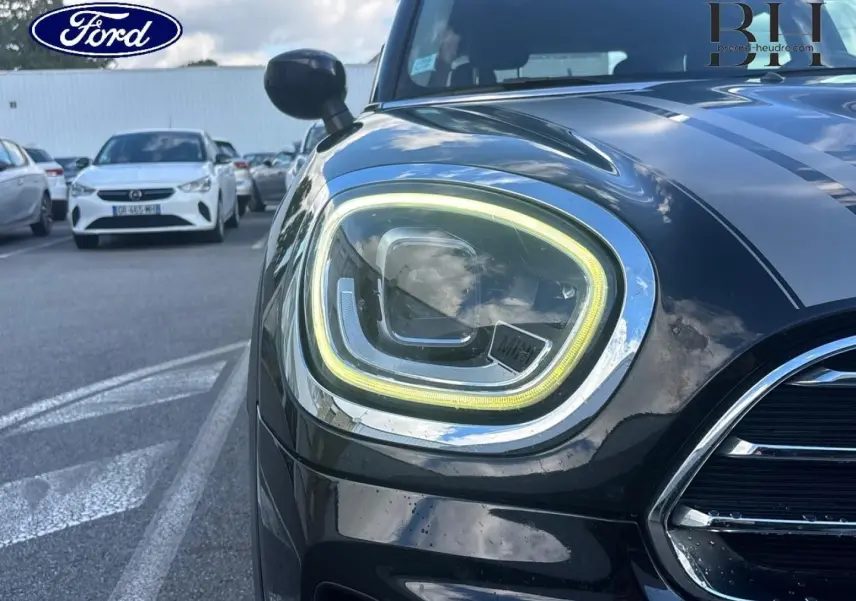 Gros plan sur le phare avant gauche allumé d’une MINI Countryman Cooper D noire avec bandes grises sur le capot.