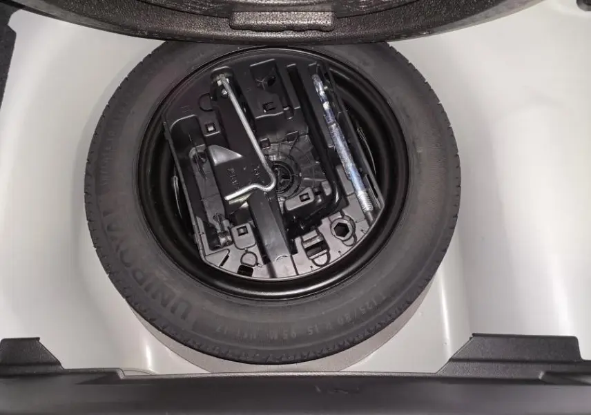 Vue plongeante sur la roue de secours galette noire et ses outils dans le coffre blanc d'une Peugeot 208 1.2i Hybrid 2025.