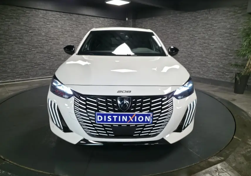 Vue frontale d'une Peugeot 208 blanche avec calandre noire et feux LED en forme de griffes allumés.