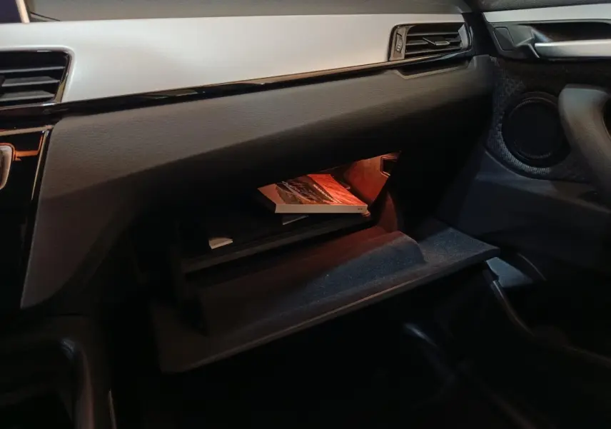 Intérieur BMW X1 sDrive18i 2021, vue côté passager sur boîte à gants ouverte avec éclairage orange et tableau de bord gris foncé.