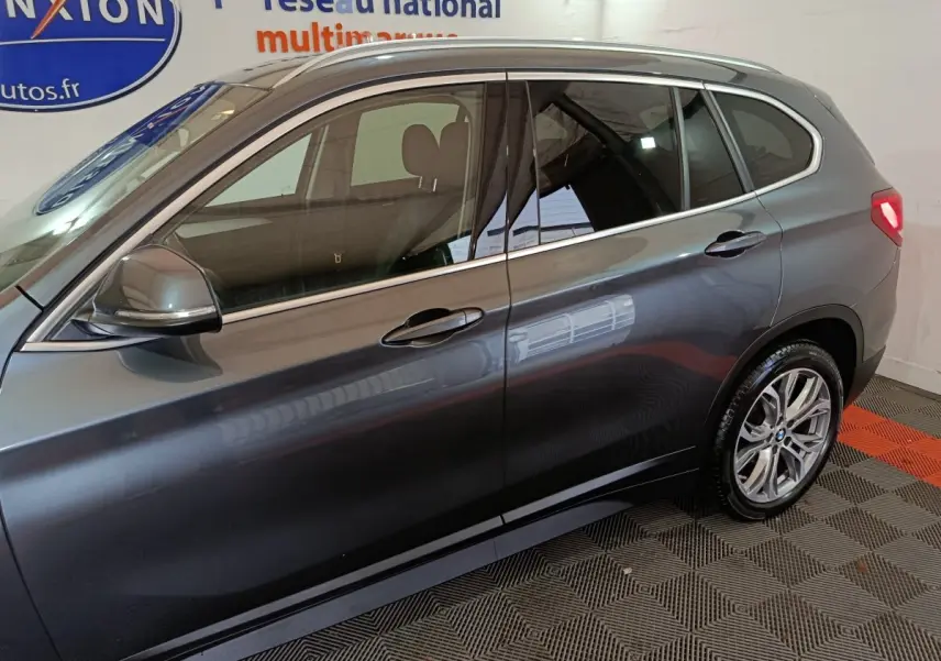 Vue latérale droite d'un BMW X1 sDrive18i Lounge gris foncé, avec jantes alliage et rails de toit noirs visibles.