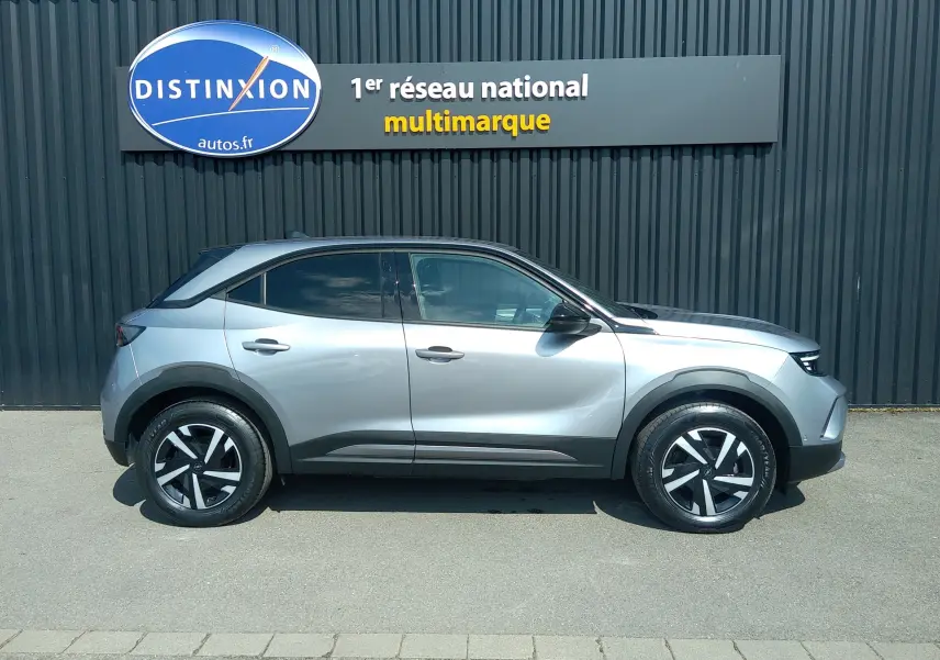 Vue de profil côté gauche d’un Opel Mokka Electric gris 2025, SUV compact avec jantes noires et grises, garé devant un mur sombre.