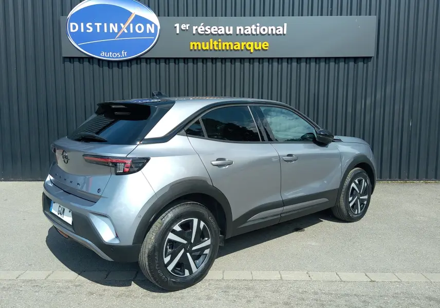Opel Mokka Electric gris en 3/4 arrière droit, avec toit noir et jantes distinctives, stationné devant un mur gris.