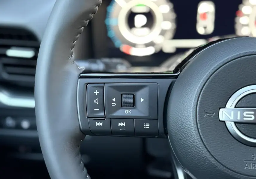 Gros plan sur les commandes gauche du volant cuir du Nissan Qashqai gris Ceramic, avec tableau de bord numérique flou en arrière-plan.