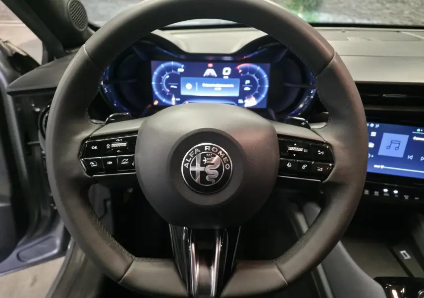 Vue rapprochée du volant noir multifonctions d'une Alfa Romeo Junior 1.2 Ibrida avec tableau de bord digital bleu.