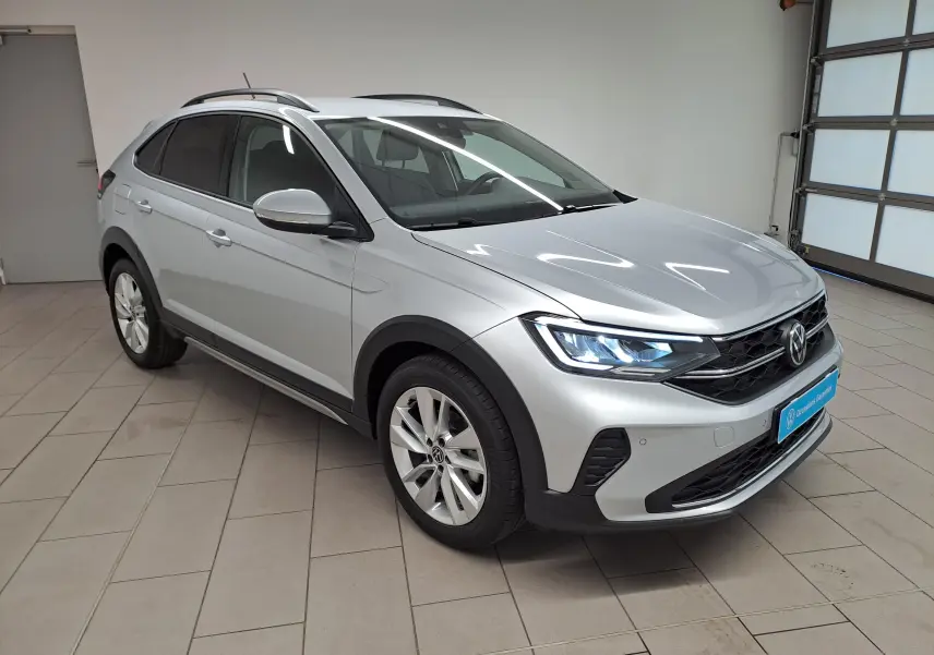 Volkswagen Taigo 2025 en Reflet d'argent, vue 3/4 avant droit avec phares allumés dans un garage.