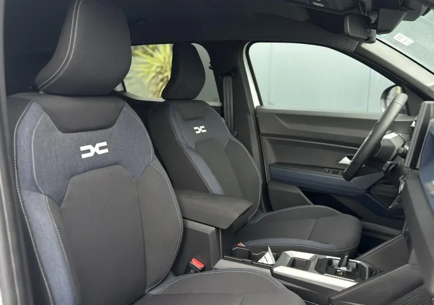 Intérieur côté conducteur du Dacia Duster blanc Glacier 2025, sièges noirs avec logo DC et accoudoir central.