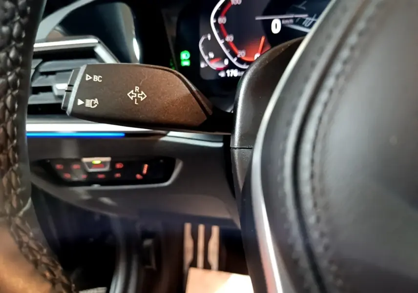 Gros plan sur la manette de clignotant à gauche du volant cuir d'une BMW Série 3 318i noire, tableau de bord numérique en arrière-plan.