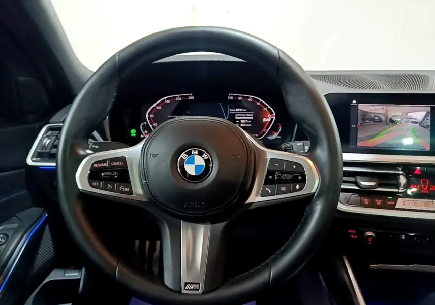 Vue rapprochée du volant cuir noir M Sport de la BMW Série 3 318i avec tableau de bord digital et écran de caméra de recul.