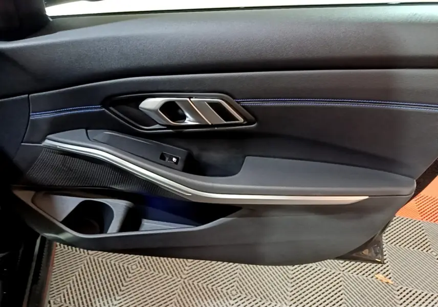 Gros plan sur la porte côté gauche noire de la BMW Série 3 318i M Sport 2022 avec surpiqûres bleues et poignée alu.