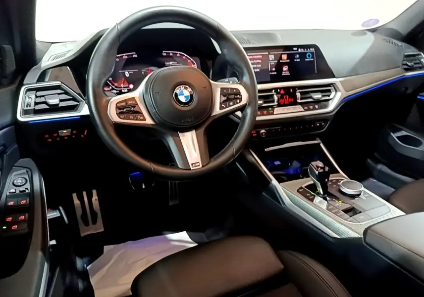 Vue intérieure avant de la BMW Série 3 318i M Sport 2022, volant cuir M, tableau de bord digital et éclairage d'ambiance bleu.