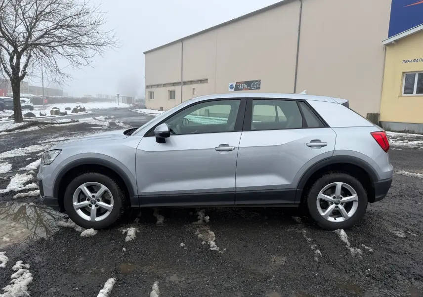 Audi Q2 gris aluminium vue de profil côté gauche sur un sol humide avec traces de neige et bâtiment en arrière-plan.