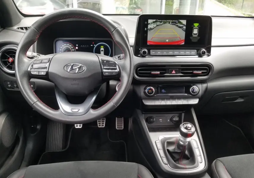 Vue intérieure avant du Hyundai Kona rouge 2023, volant cuir, écran tactile avec caméra de recul et boîte manuelle.