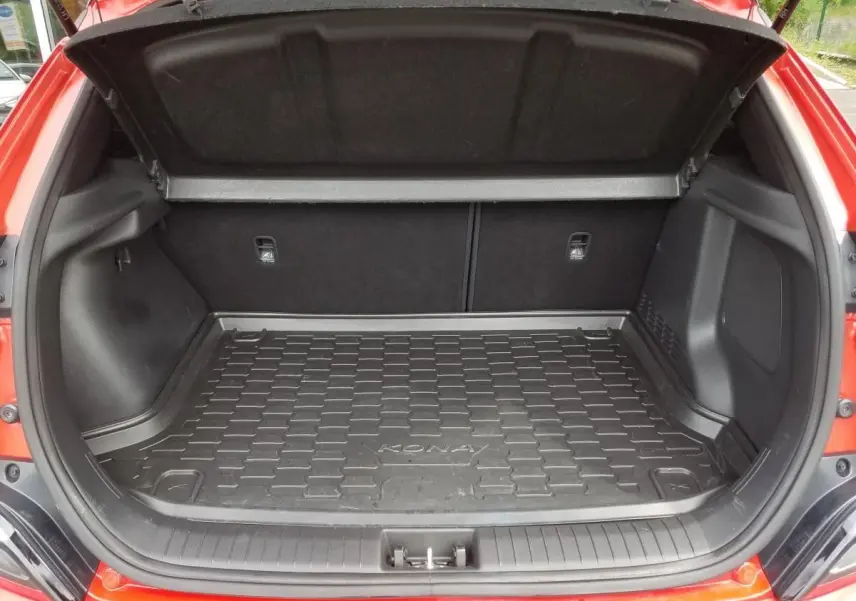 Coffre ouvert vu de l'arrière d'un Hyundai Kona rouge avec tapis de coffre noir marqué Kona.