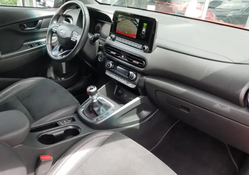 Vue intérieure côté conducteur du Hyundai Kona rouge 2023, avec tableau de bord numérique et levier de vitesse manuel.
