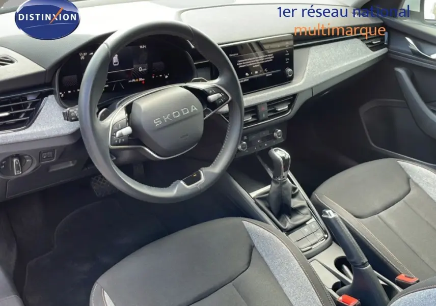 Vue intérieure centrée sur le volant et la console centrale d'une Skoda Scala blanche Glacier avec sellerie tissu gris noir.
