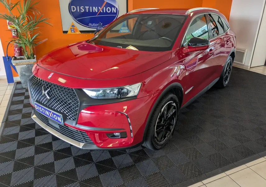 Vue 3/4 avant d’un DS7 rouge 2019 avec attelage, jantes noires et calandre distinctive dans un showroom.