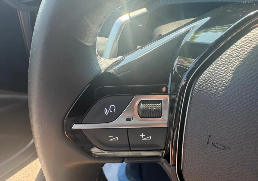 Gros plan sur les commandes gauche du volant noir du Peugeot 2008 Hybrid 145 gris, avec molette et boutons de volume.
