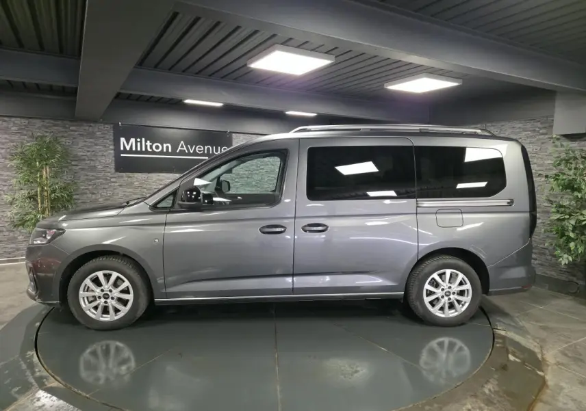 Vue de profil côté gauche d’un Ford Tourneo Connect gris métallisé avec barres de toit argentées en intérieur showroom.