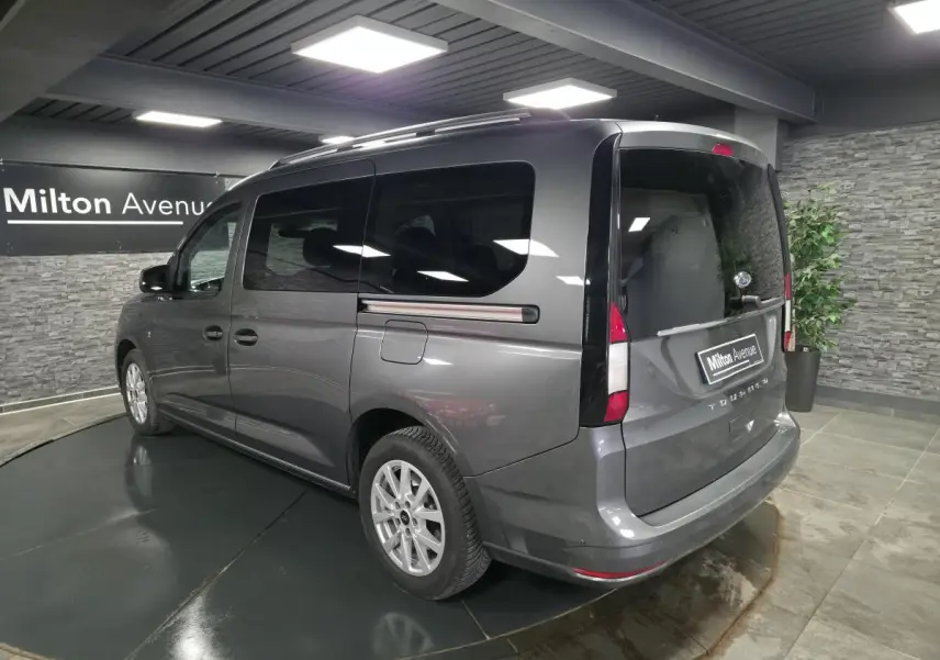 Vue 3/4 arrière droit du Ford Transit Connect Tourneo gris métallisé avec barres de toit argentées en intérieur showroom.