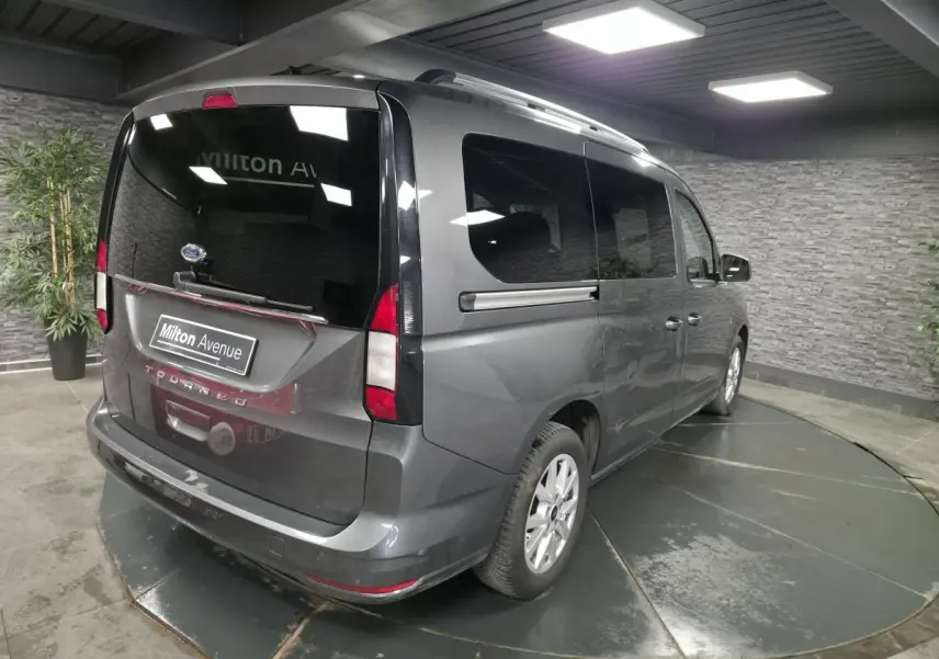 Vue 3/4 arrière droite du Ford Tourneo Connect gris métallisé avec vitres teintées et barres de toit argentées.