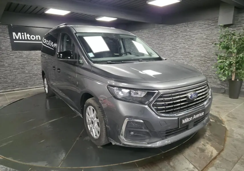 Vue 3/4 avant droit du Ford Transit Connect gris métallisé avec calandre noire et barres de toit argentées en showroom.