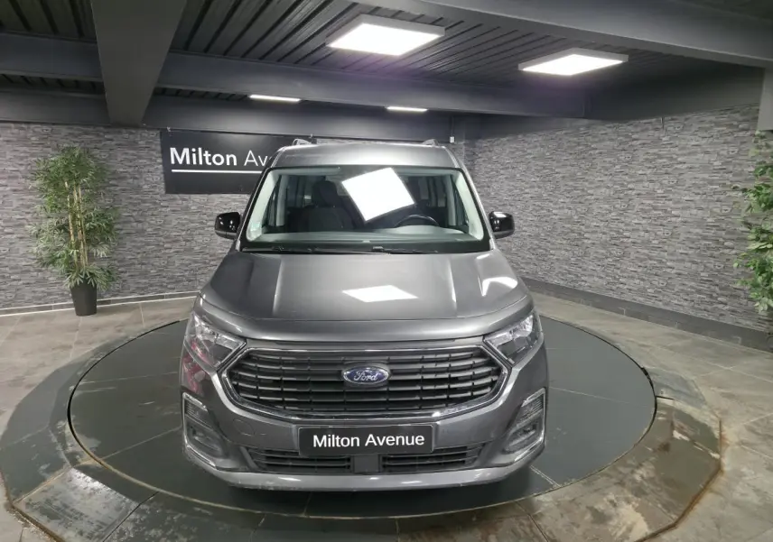Vue avant d'un Ford Transit Connect gris métallisé avec calandre noire et barres de toit argentées en showroom.