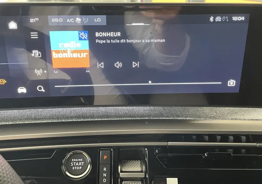 Vue intérieure du tableau de bord du Peugeot 3008 Hybrid 2026, avec écran tactile affichant une radio et bouton start/stop visible.