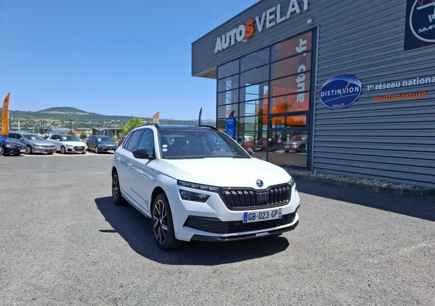 Vue 3/4 avant d'un Skoda Kamiq blanc 2021 avec jantes alliage noires devant un bâtiment commercial.