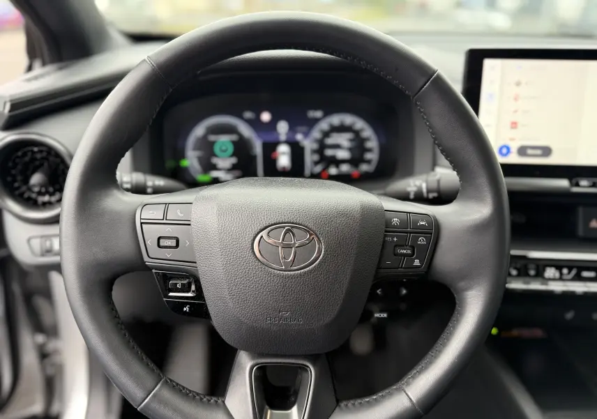 Vue rapprochée du volant noir du Toyota C-HR Hybride 2024 avec commandes intégrées et tableau de bord numérique flou.