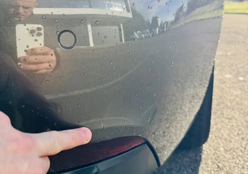 Gros plan sur l'aile arrière droite gris amazonite d'une Peugeot 508, avec un doigt pointant une rayure près du feu arrière.