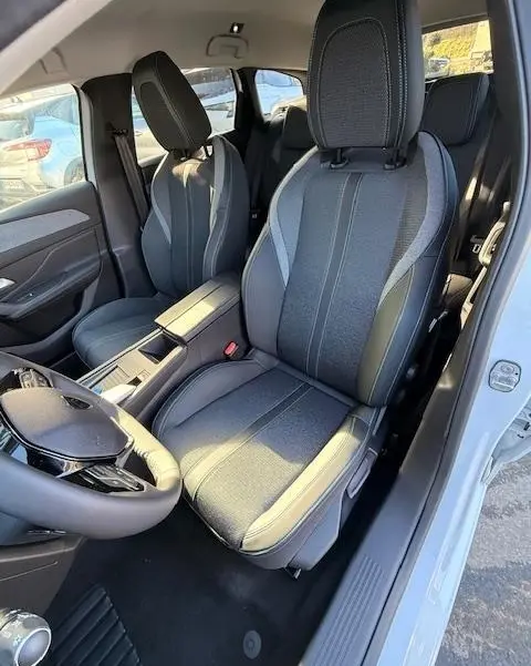 Vue intérieure côté conducteur de la Peugeot 308 Hybrid 145 e-DCS6 Allure 2025 blanche, sièges tissu gris avec surpiqûres.