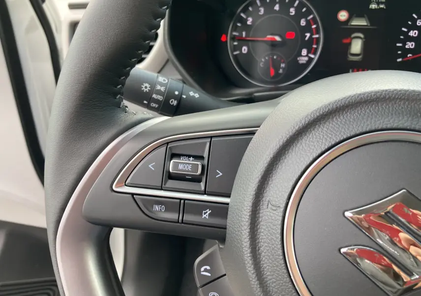 Gros plan sur le volant cuir multifonctions gris d’une Suzuki Swift blanche, avec commandes audio et tableau de bord en arrière-plan.