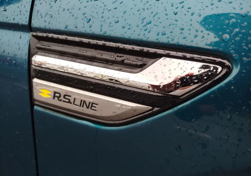 Gros plan sur l'emblème R.S. Line chromé sur la portière bleu Zanzibar d'une Renault Arkana 2022, avec gouttes de pluie.