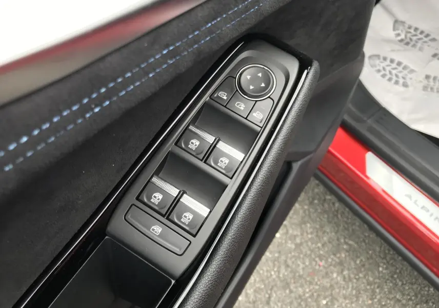 Gros plan sur les commandes de lève-vitres côté conducteur avec surpiqûres bleues et seuil de porte rouge Alpine visible.