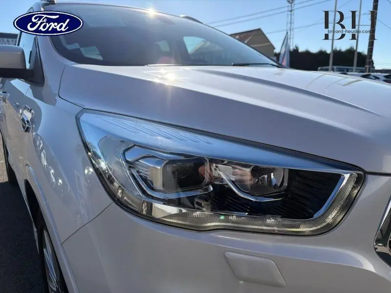 Gros plan sur le phare avant droit du Ford Kuga blanc Premium Platinum sous un ciel ensoleillé.