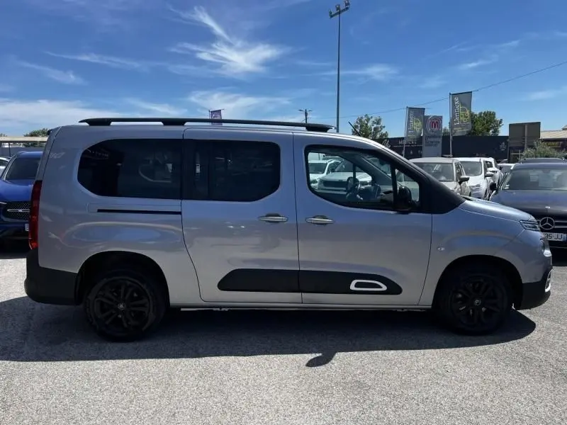 Vue de profil côté gauche d'un Citroën Berlingo XL gris métal avec protections noires et jantes foncées, stationné en extérieur.