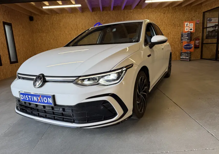 Volkswagen Golf 2.0 TDI 150 DSG7 R-Line blanc vue 3/4 avant en intérieur avec jantes noires et phares allumés.