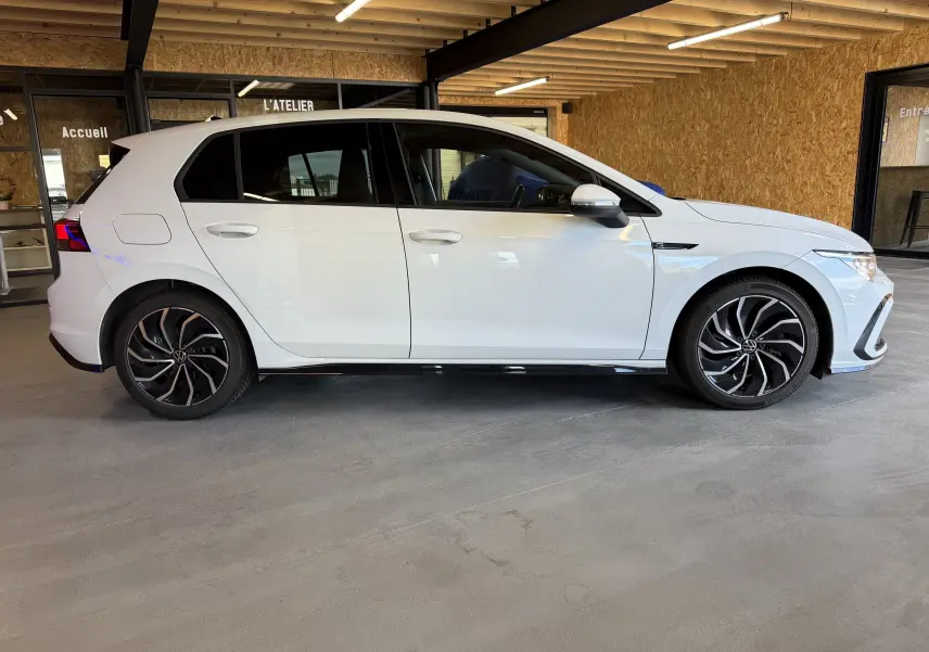 Vue de profil côté gauche d'une Volkswagen Golf 2.0 TDI 150 DSG7 R-Line blanche avec jantes noires et détails sportifs.