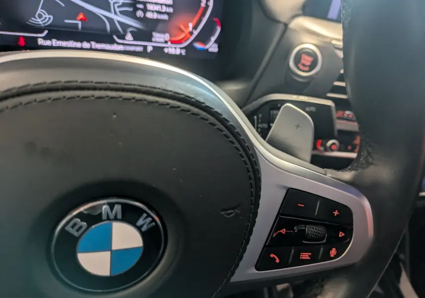 Gros plan sur le volant noir de la BMW X4 blanc, avec commandes intégrées et tableau de bord numérique en arrière-plan.