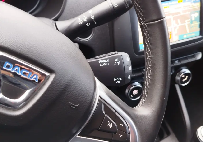 Gros plan sur le volant noir du Dacia Duster rouge Fusion 2020, avec commandes audio et écran tactile en arrière-plan.