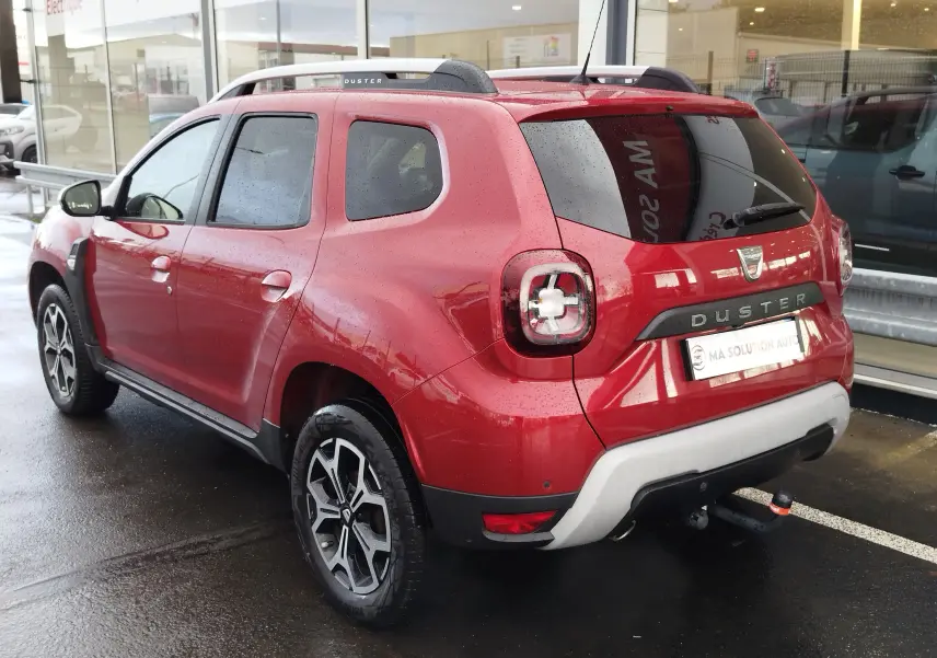 Vue 3/4 arrière droite du Dacia Duster rouge fusion 2020 avec barres de toit et attelage visible.