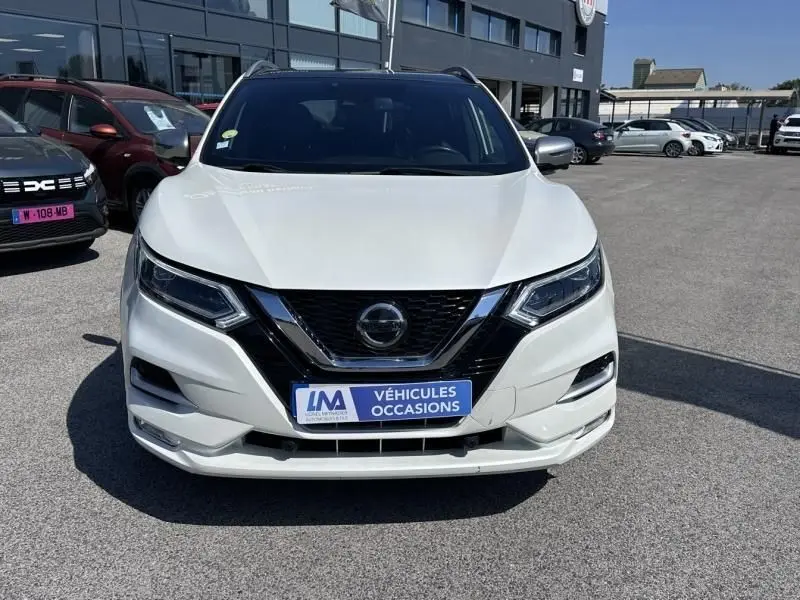 Vue avant d'un Nissan Qashqai blanc 2019 avec calandre noire et badge central, stationné devant un bâtiment commercial.