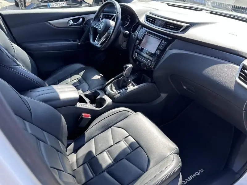 Vue intérieure côté conducteur du Nissan Qashqai blanc 2019, avec sièges cuir noirs et console centrale équipée d'un écran tactile.