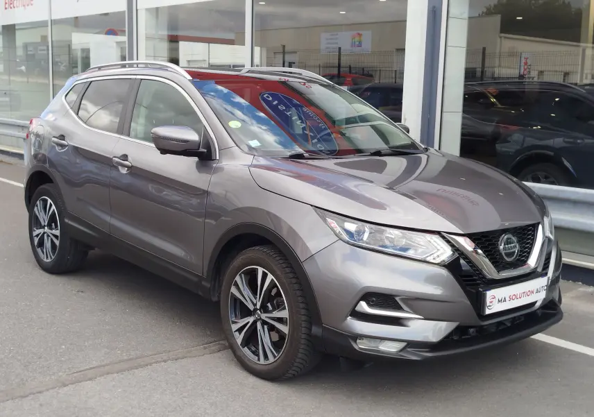 Nissan Qashqai gris Squale en 3/4 avant droit, avec jantes alliage et calandre chromée visible devant un showroom.