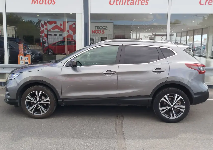 Vue de profil côté gauche d'un Nissan Qashqai gris Squale avec jantes alliage et vitres teintées, stationné devant un showroom.