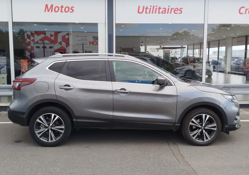 Vue de profil côté gauche d'un Nissan Qashqai gris Squale avec jantes alliage et vitres teintées, stationné devant un showroom.